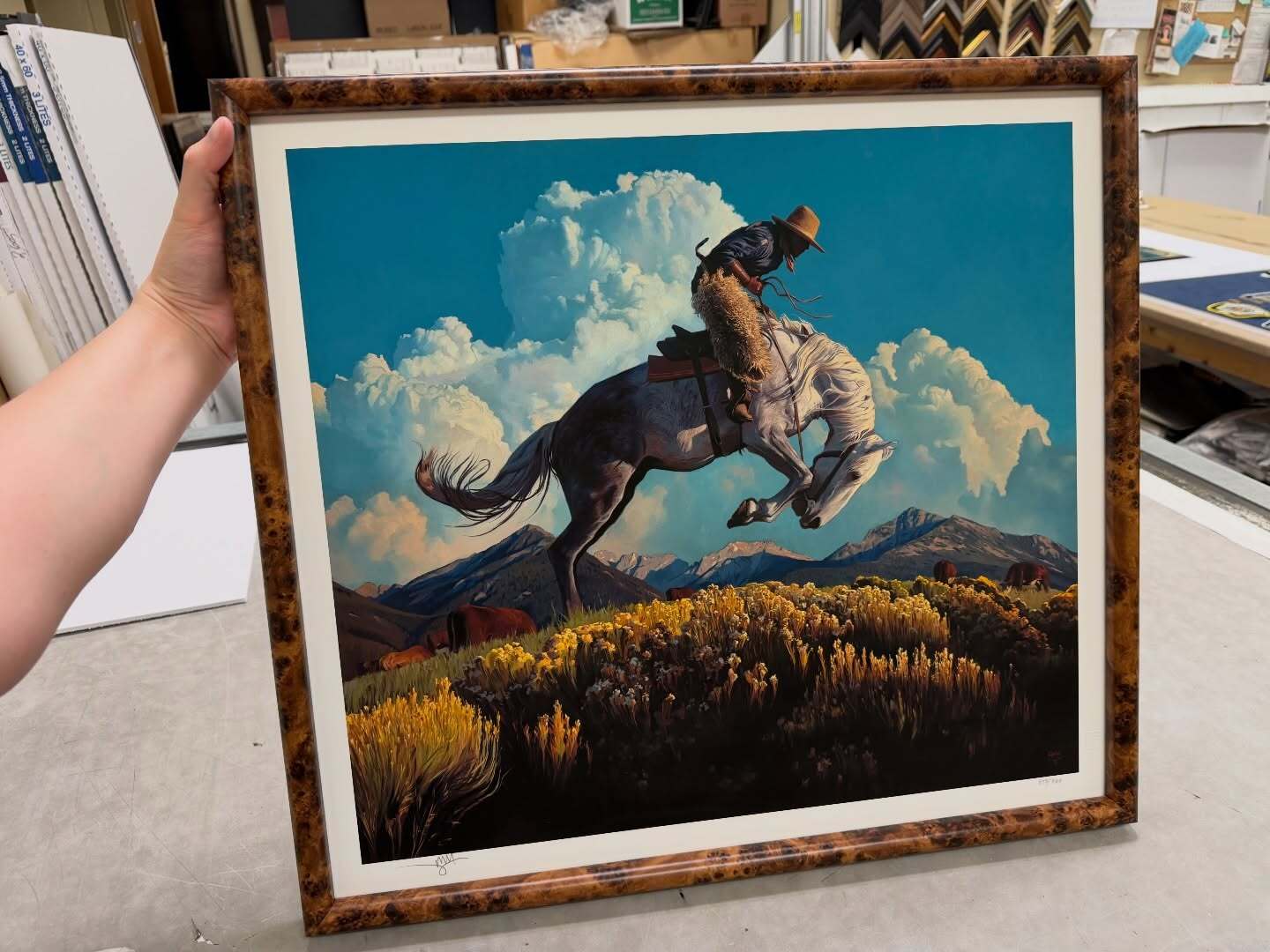 A framed art print depicting a cowboy riding a bucking horse over a grassy field and mountains, held up by a person's hand in what appears to be an art store or gallery.