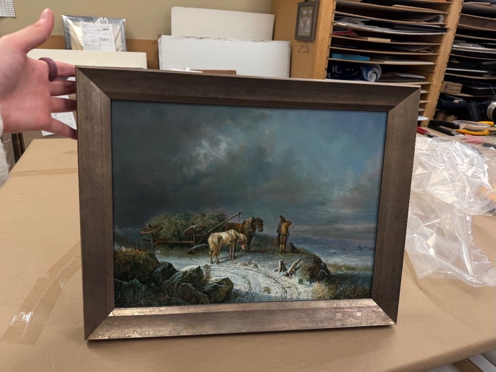 A framed classic oil painting depicting a farmer, two horses, and a wagon on a snowy trail under a dramatic sky.