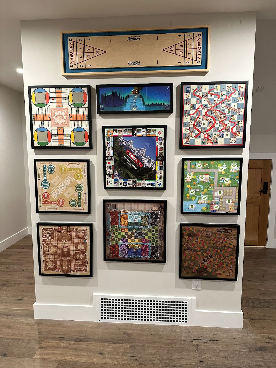 A wall display of framed classic board games including Monopoly, Chutes and Ladders, Scrabble, and others, arranged in a grid pattern above a floor vent.