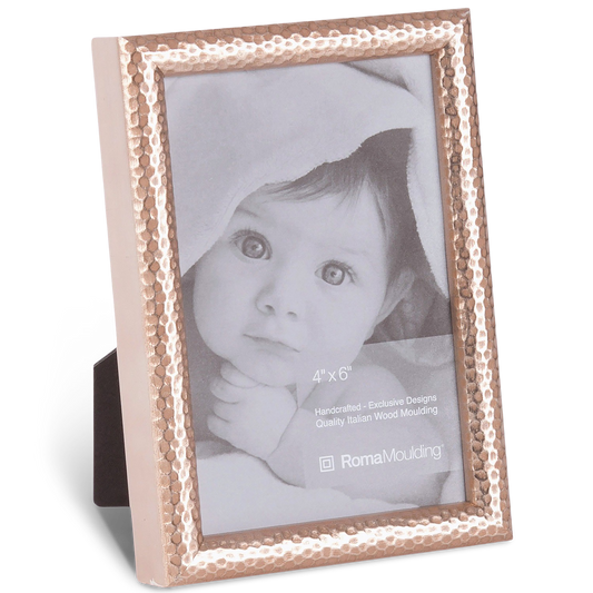 A hammered metal photo frame holds a black-and-white image of an infant with wide eyes, partially covered by a blanket, conveying innocence and warmth.