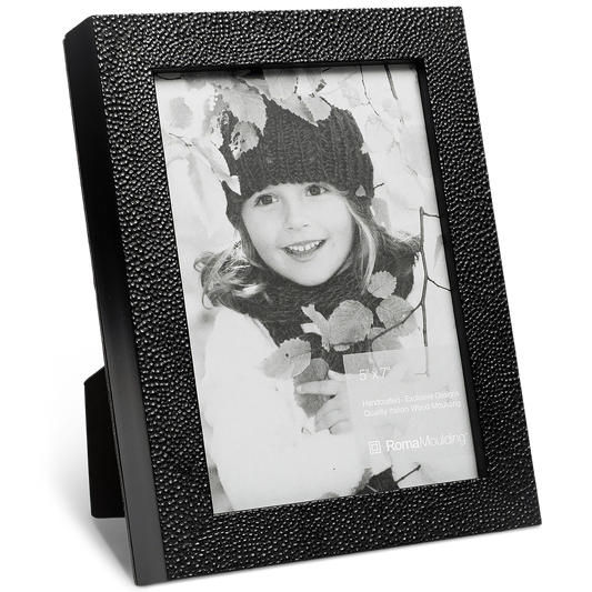 Black and white photo of a smiling child in a knit hat, surrounded by leaves, in a textured black picture frame. Warm, nostalgic tone.