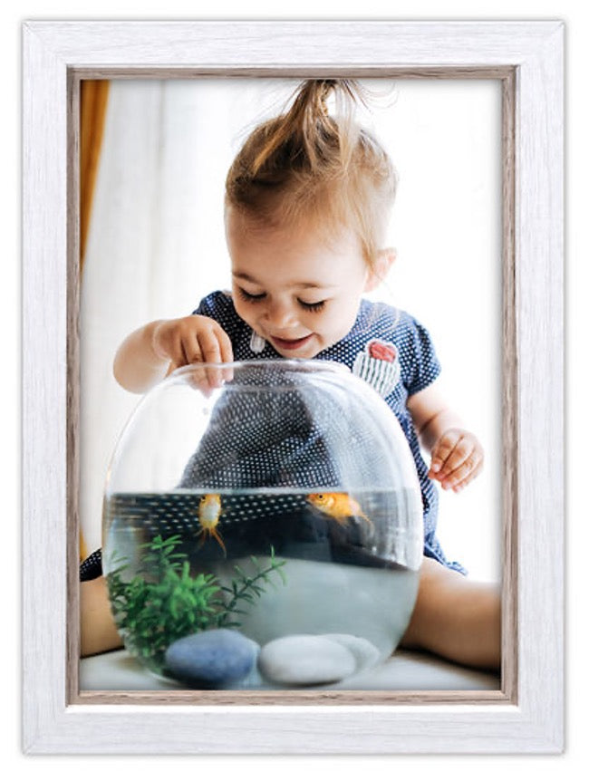 A smiling toddler in a polka dot dress sits with a glass fishbowl, curiously watching two goldfish swim. The scene is playful and joyful.