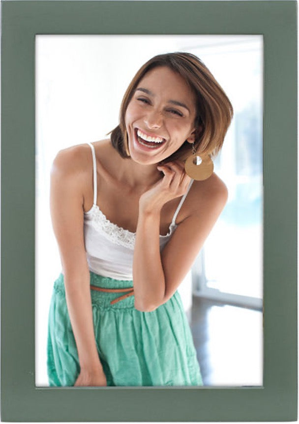 Smiling woman in white tank top and green skirt, holding her earring. Bright, airy background with a joyful, lively atmosphere.