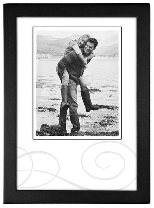 A joyful black-and-white photo of a woman playfully piggybacking on a man at the beach. Both are smiling, wearing boots, and the scene is framed neatly.