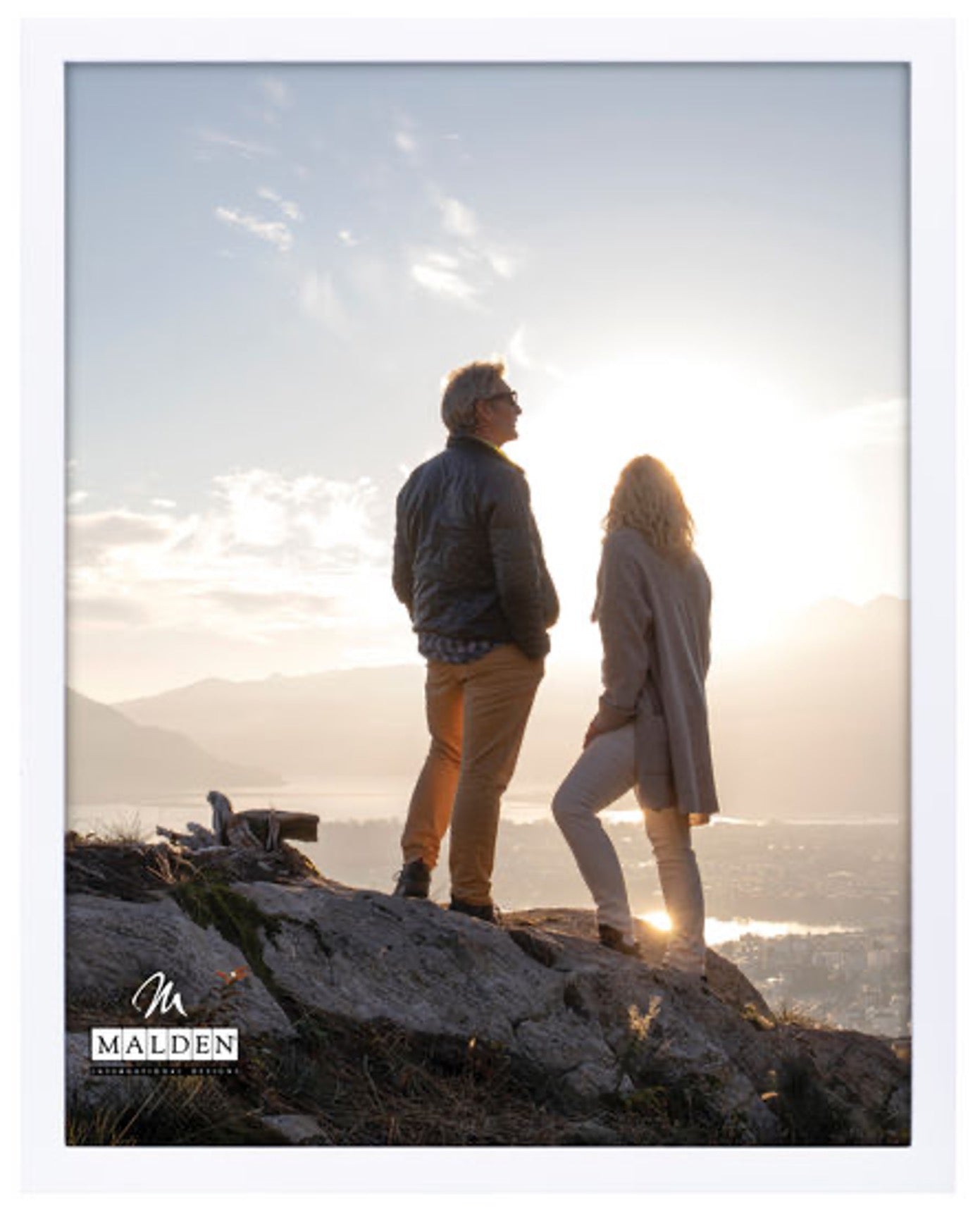 A couple stands on a rocky hilltop at sunset, overlooking a scenic valley. The scene conveys tranquility and reflection under a bright, glowing sky.
