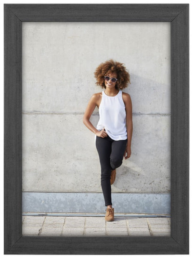 A woman with curly hair, wearing sunglasses, a white top, black pants, and brown shoes, leans casually against a concrete wall, exuding a relaxed vibe.