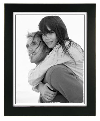 A black-and-white photo of a woman embracing a man from behind, both smiling softly. The image is set in a dark blue frame, conveying warmth and intimacy.