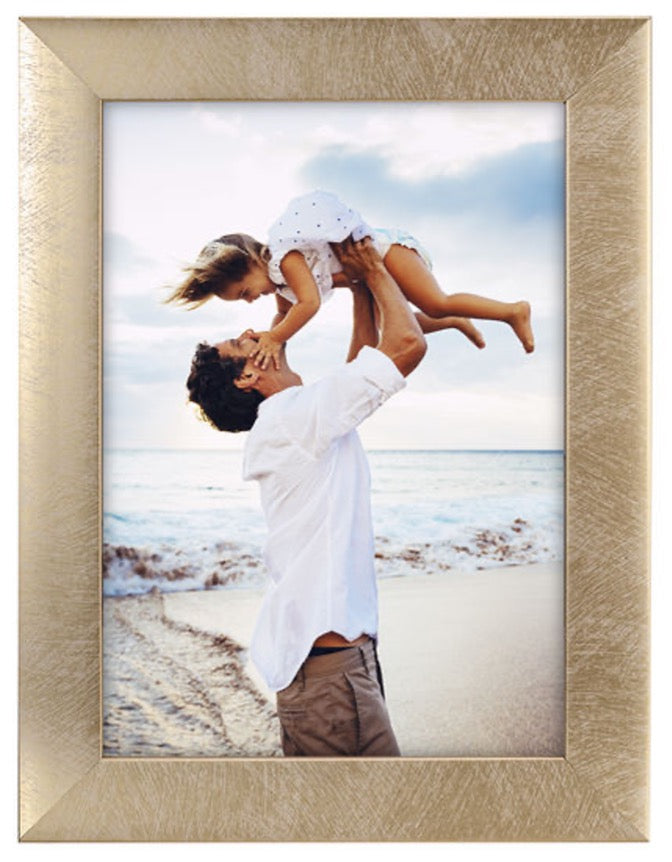 A dad joyfully lifts his daughter into the air on a beach, both smiling. The sun sets in the background, creating a warm, serene atmosphere.