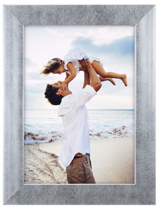 A man joyfully lifts a young child above him at a beach, both smiling. The ocean and a cloudy sky form the serene background. Framed in silver.