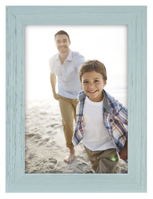 Framed photo of a joyful child in a plaid shirt running on the beach, followed by a smiling adult in casual attire, both under bright sunlight.
