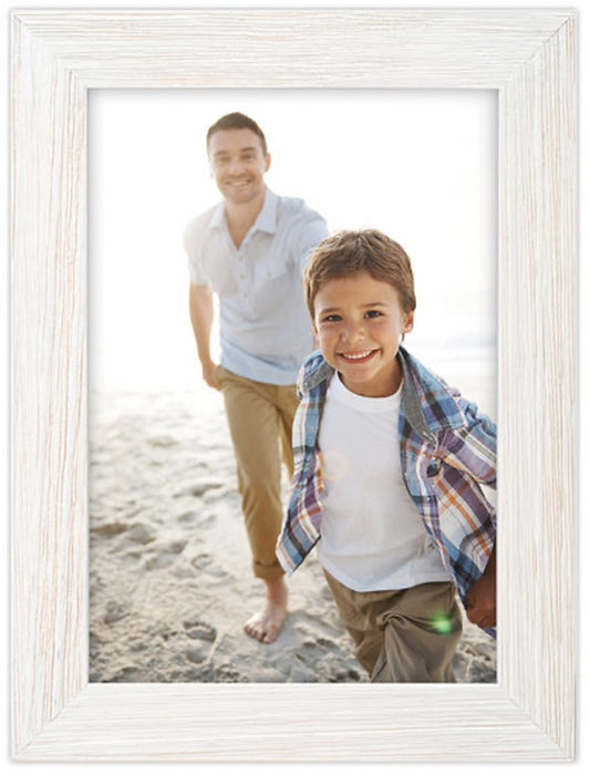 A smiling child in a plaid shirt runs toward the camera on a sunny beach, with a man happily following behind. A light wood frame surrounds the photo.