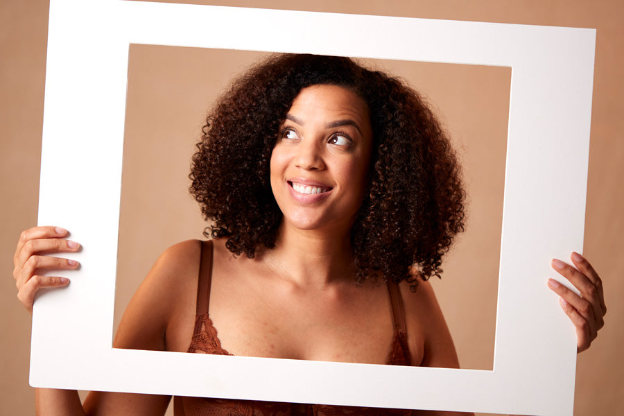 A woman with curly hair, smiling and looking upwards, holds a blank white frame. She wears a brown top against a beige background, conveying a cheerful mood.