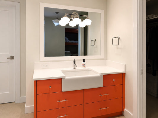 A modern bathroom vanity featuring a white farmhouse sink and white countertop, supported by a bright orange cabinet with silver handles, beneath a large white-framed mirror with a multi-light fixture.