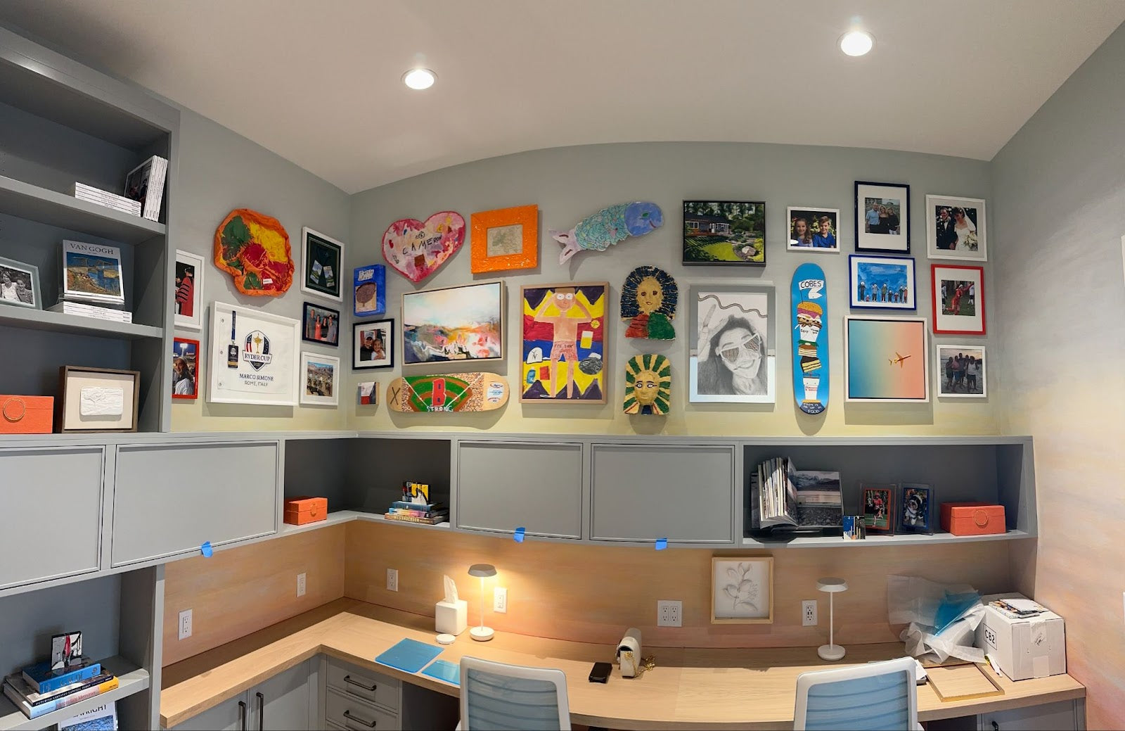 A modern home office with a large L-shaped desk, grey cabinetry, and a vibrant gallery wall featuring eclectic artwork, family photos, and a decorated skateboard.