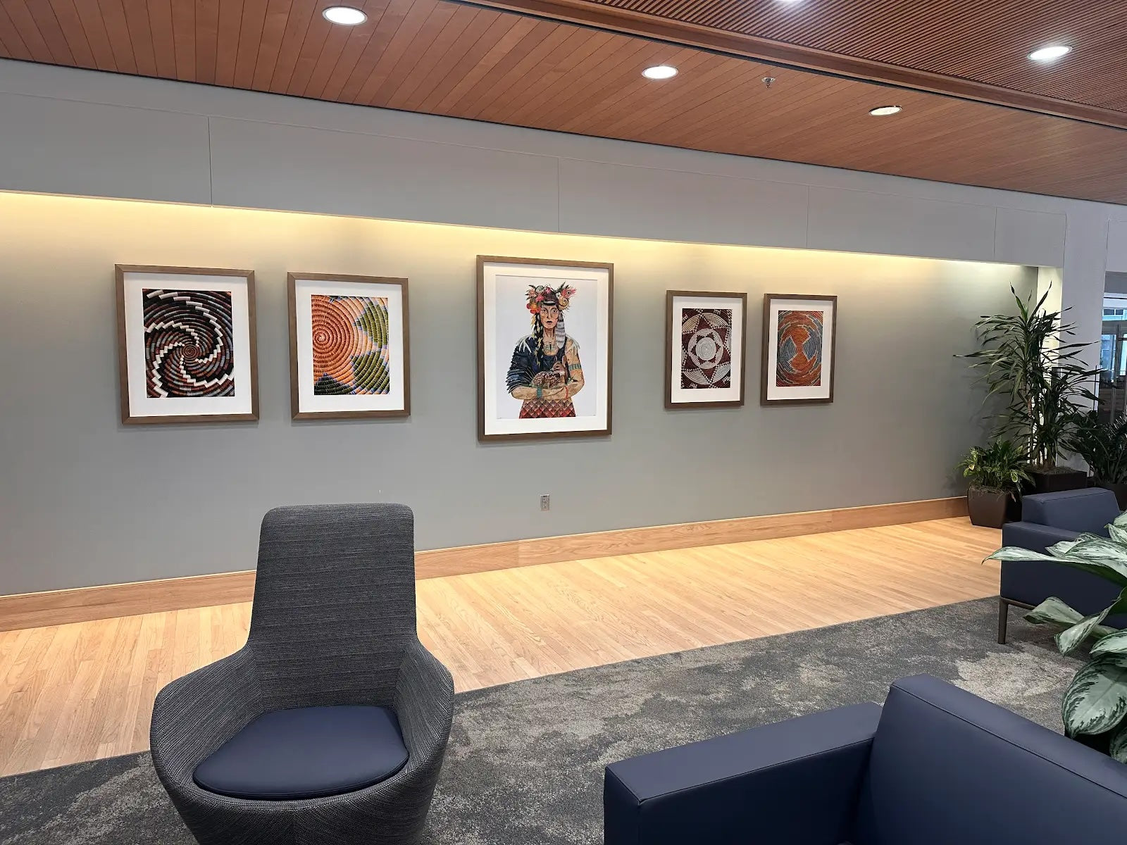 Modern lobby with wooden ceiling, framed artwork on gray wall, and blue chairs on wooden floor.
