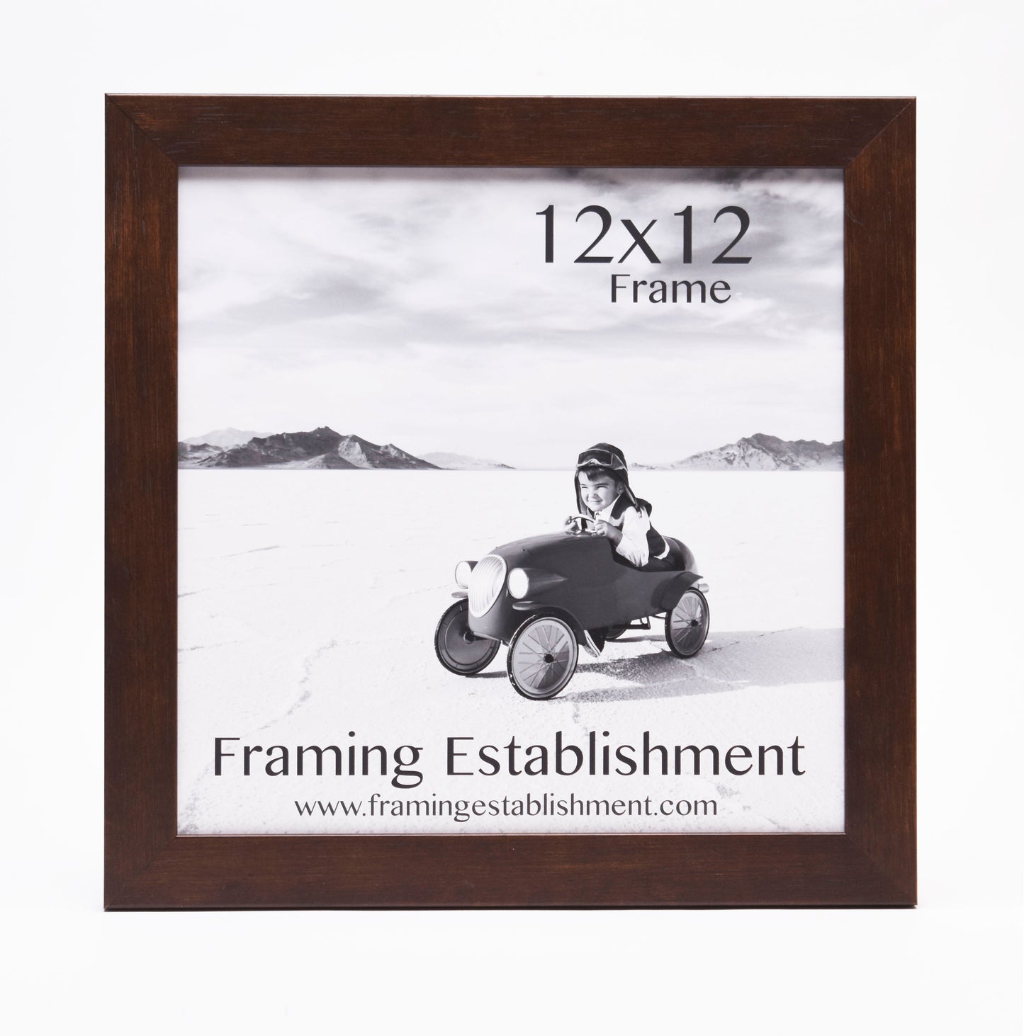 A black and white photo in a 12x12 dark wooden frame shows a smiling child in a toy car on a flat landscape. Text: Framing Establishment.