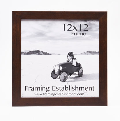 A black and white photo in a 12x12 dark wooden frame shows a smiling child in a toy car on a flat landscape. Text: Framing Establishment.