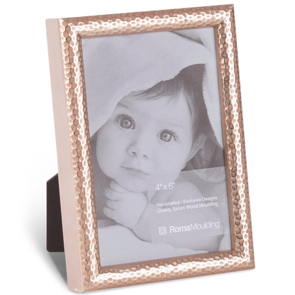 A hammered metal photo frame holds a black-and-white image of an infant with wide eyes, partially covered by a blanket, conveying innocence and warmth.