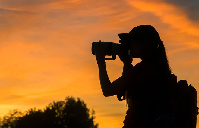 Silhouette of a person taking a photo with a camera against an orange sunset sky.