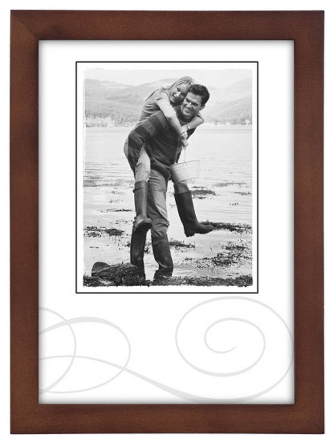 A black-and-white photo in a brown frame shows a smiling couple outdoors. The woman playfully rides on the man's back near a scenic lake shore.