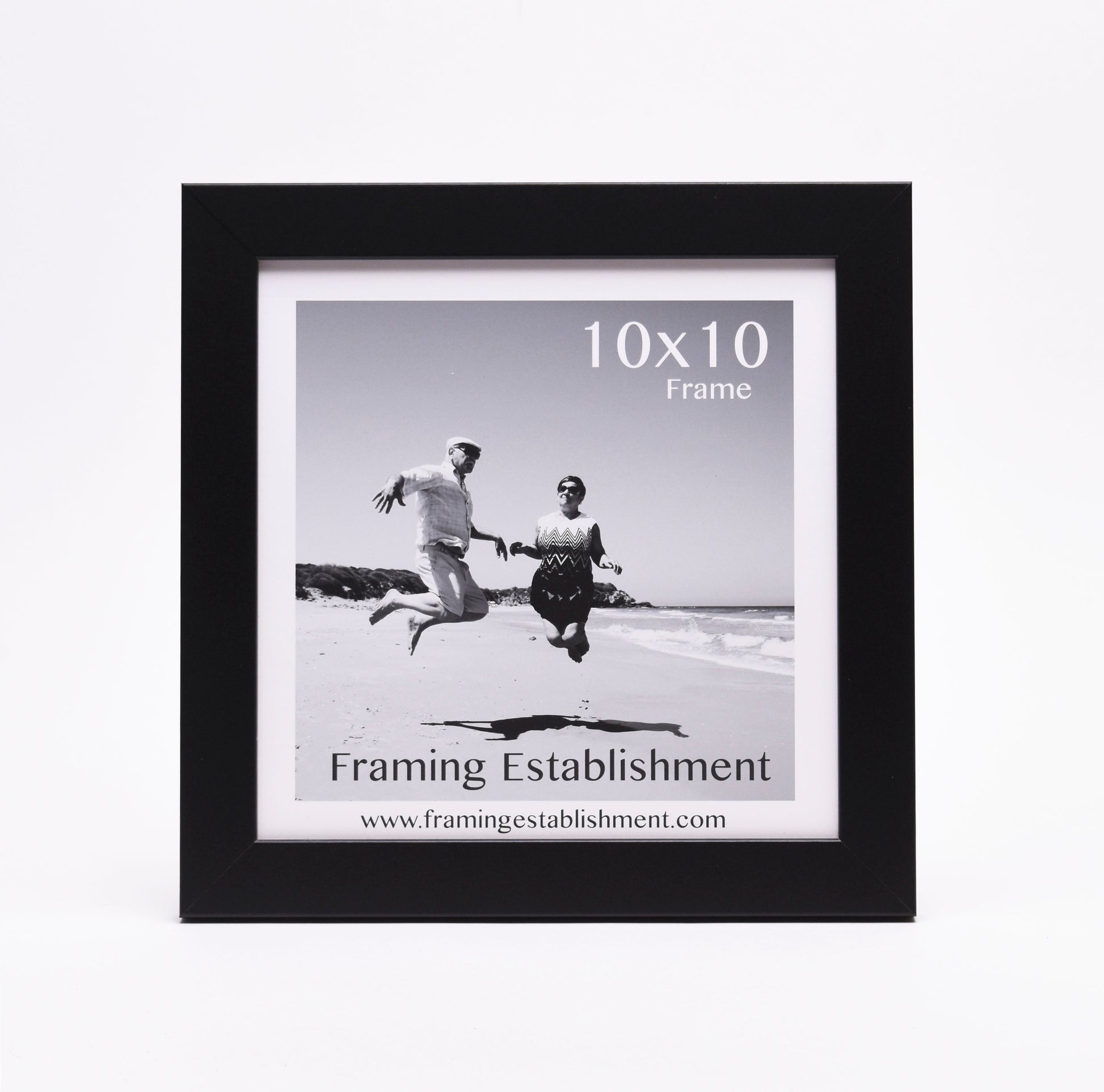 Black frame around a black-and-white photo of a man and woman joyfully jumping on a beach. "10x10 Frame" text and "Framing Establishment" logo visible.