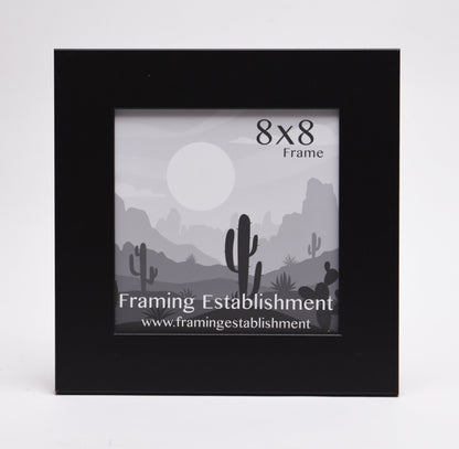 A black 8x8 photo frame with a minimalist desert landscape illustration inside. The image features cacti, hills, and a large sun. A calm, serene tone.