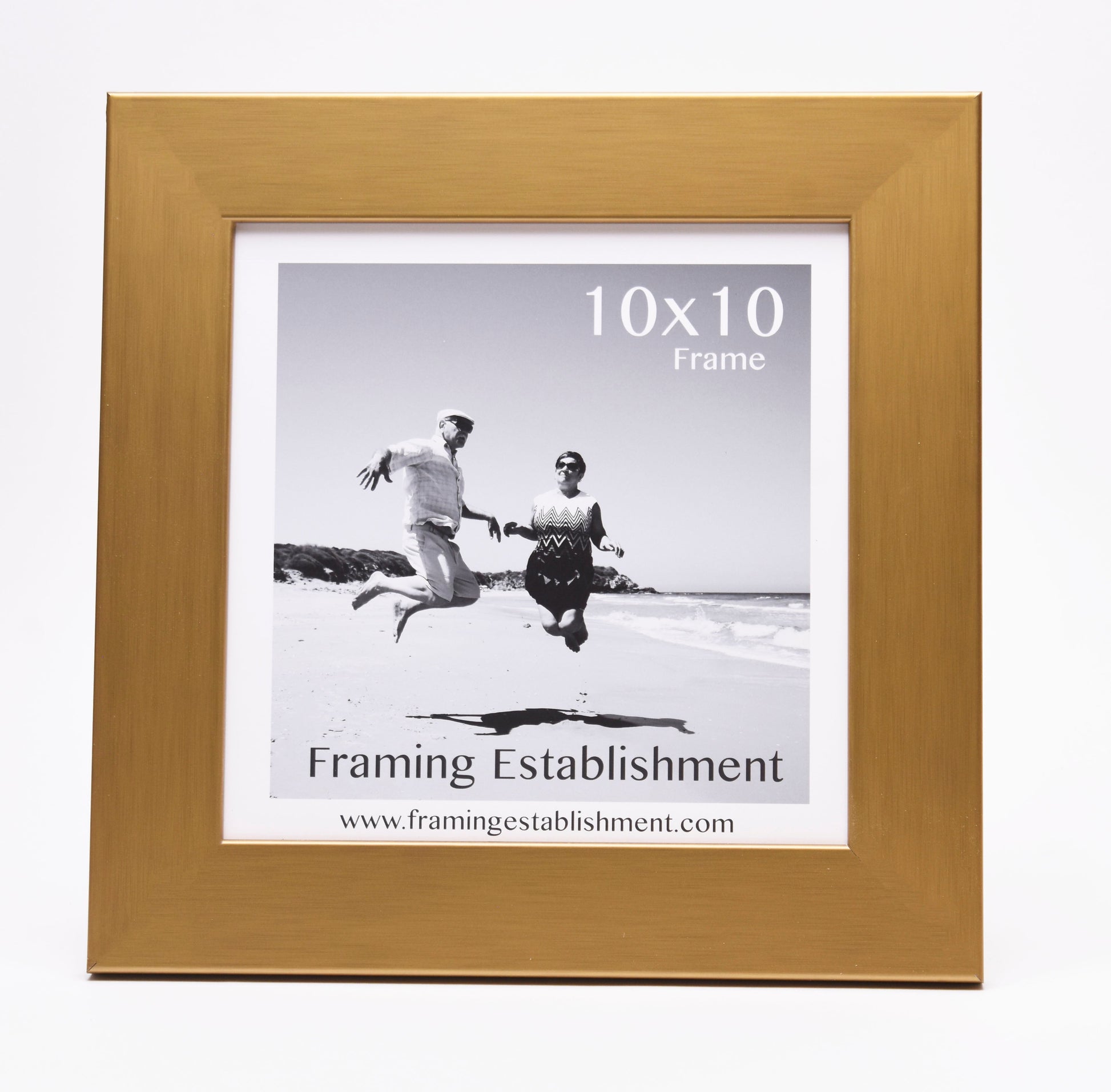 A gold frame displaying a black and white photo of two people joyfully jumping on a beach. Text reads "10x10 Frame" and "Framing Establishment."