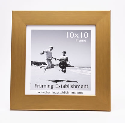 A gold frame displaying a black and white photo of two people joyfully jumping on a beach. Text reads "10x10 Frame" and "Framing Establishment."
