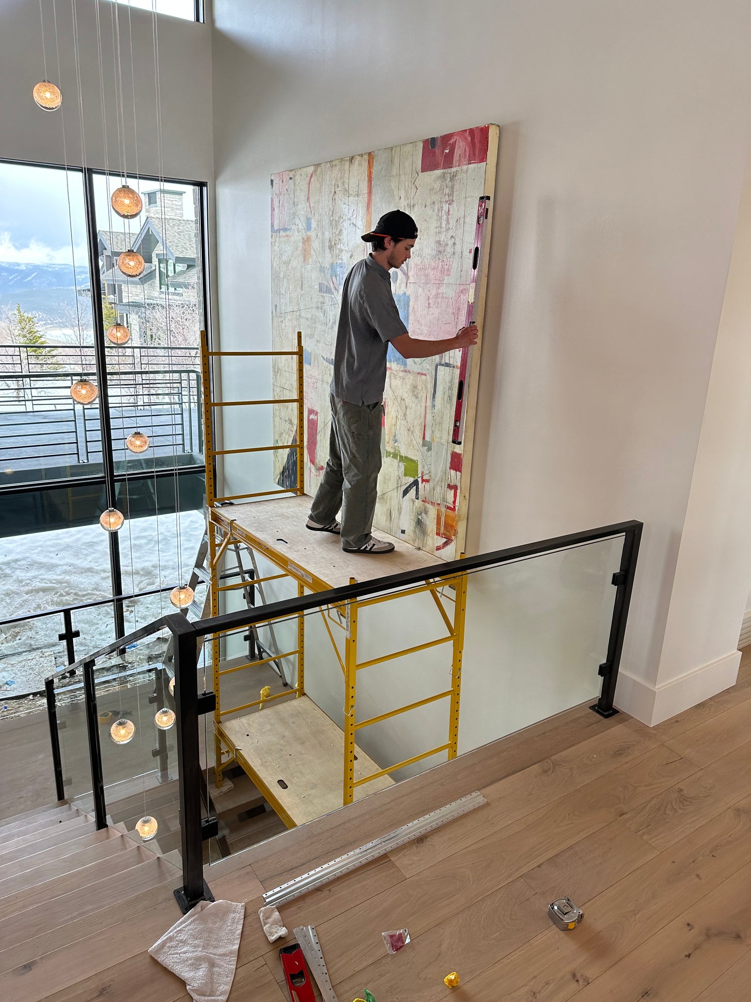 Person working on a ceiling with tools and materials in a room with wooden floor and large window.