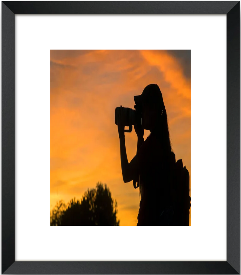 Silhouette of a person taking a photo against an orange sunset sky