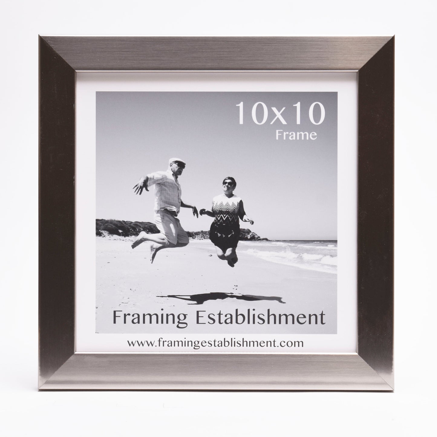 Black and white photo of a joyful couple jumping on a sunny beach. They are framed in a stylish silver 10x10 frame from Framing Establishment.