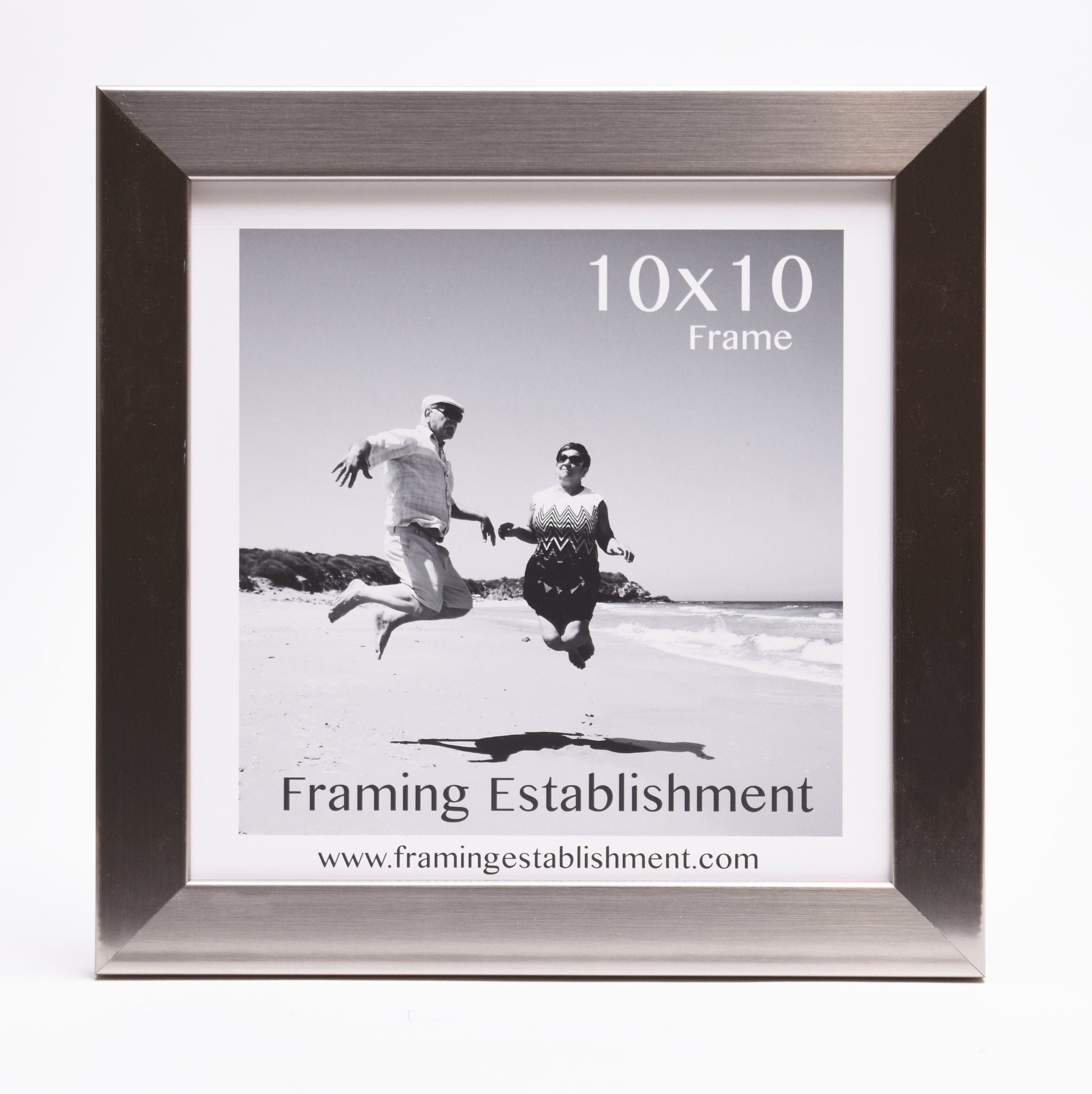 Black and white photo of a joyful couple jumping on a sunny beach. They are framed in a stylish silver 10x10 frame from Framing Establishment.
