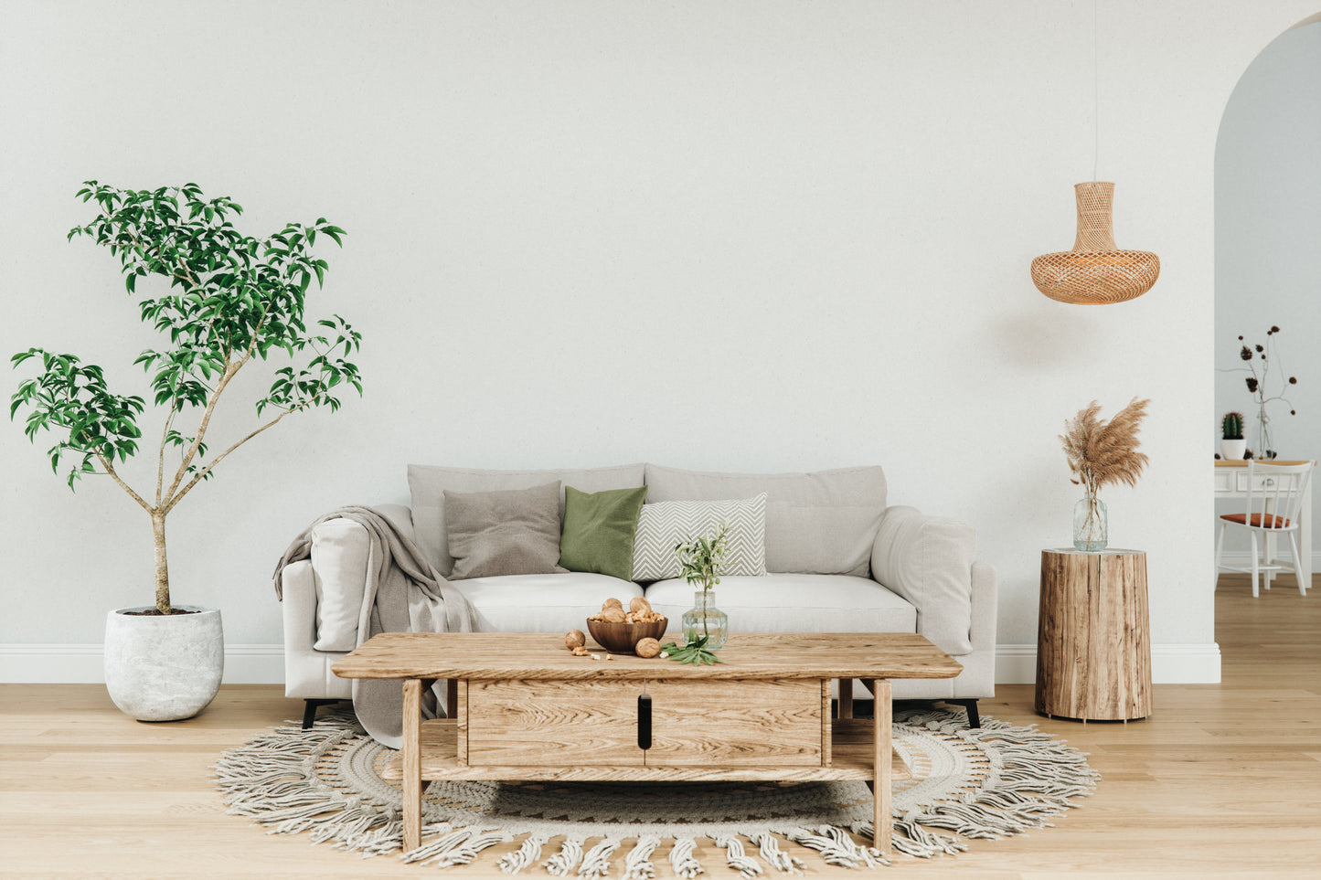 A cozy living room with a beige sofa adorned with green and gray pillows. There's a large potted plant on the left and a wooden coffee table on a round rug. A wicker pendant light hangs above, while a rustic side table with dried flowers adds warmth. The tone is tranquil and inviting.