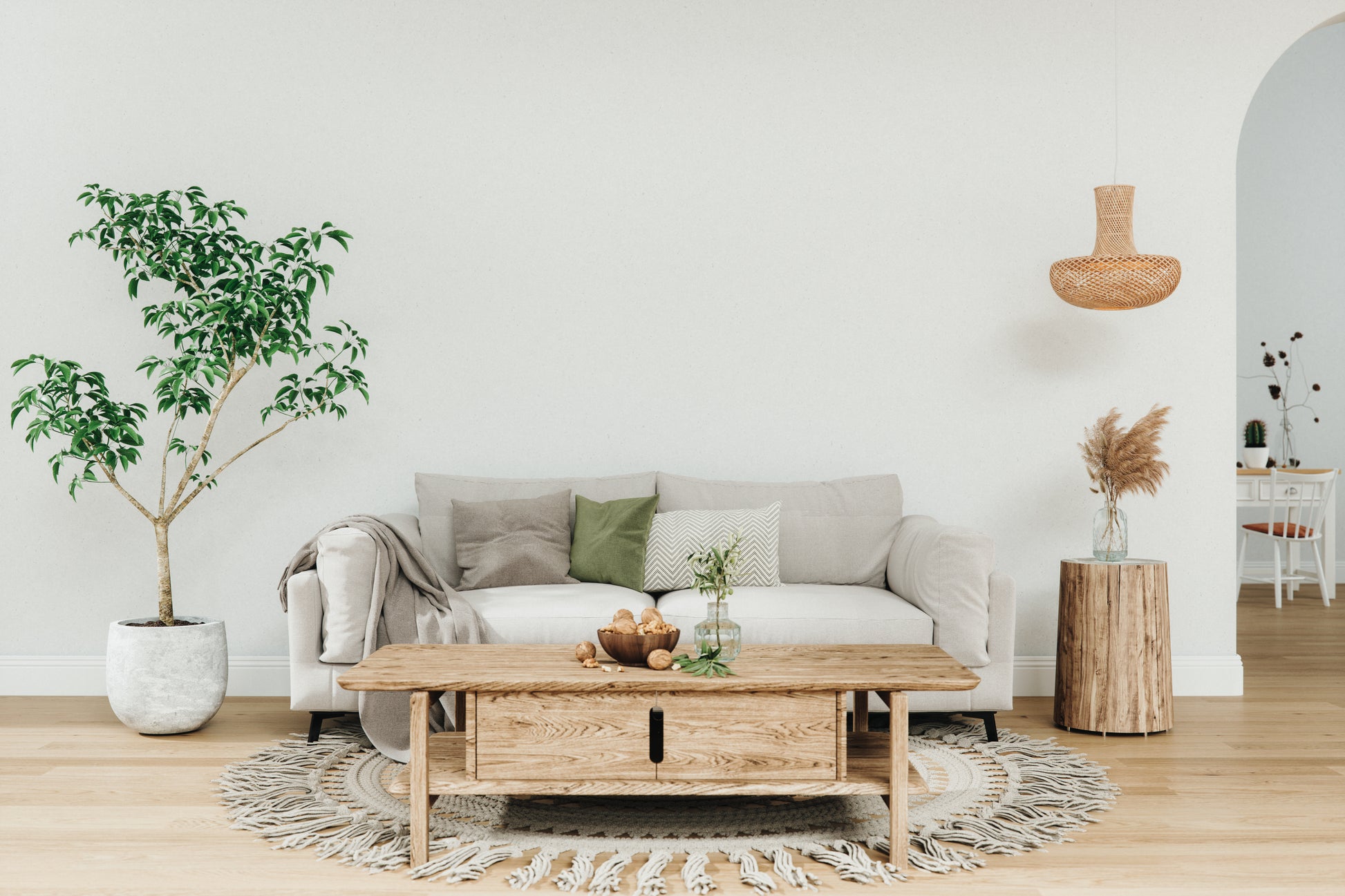 A cozy living room with a beige sofa adorned with green and gray pillows. There's a large potted plant on the left and a wooden coffee table on a round rug. A wicker pendant light hangs above, while a rustic side table with dried flowers adds warmth. The tone is tranquil and inviting.