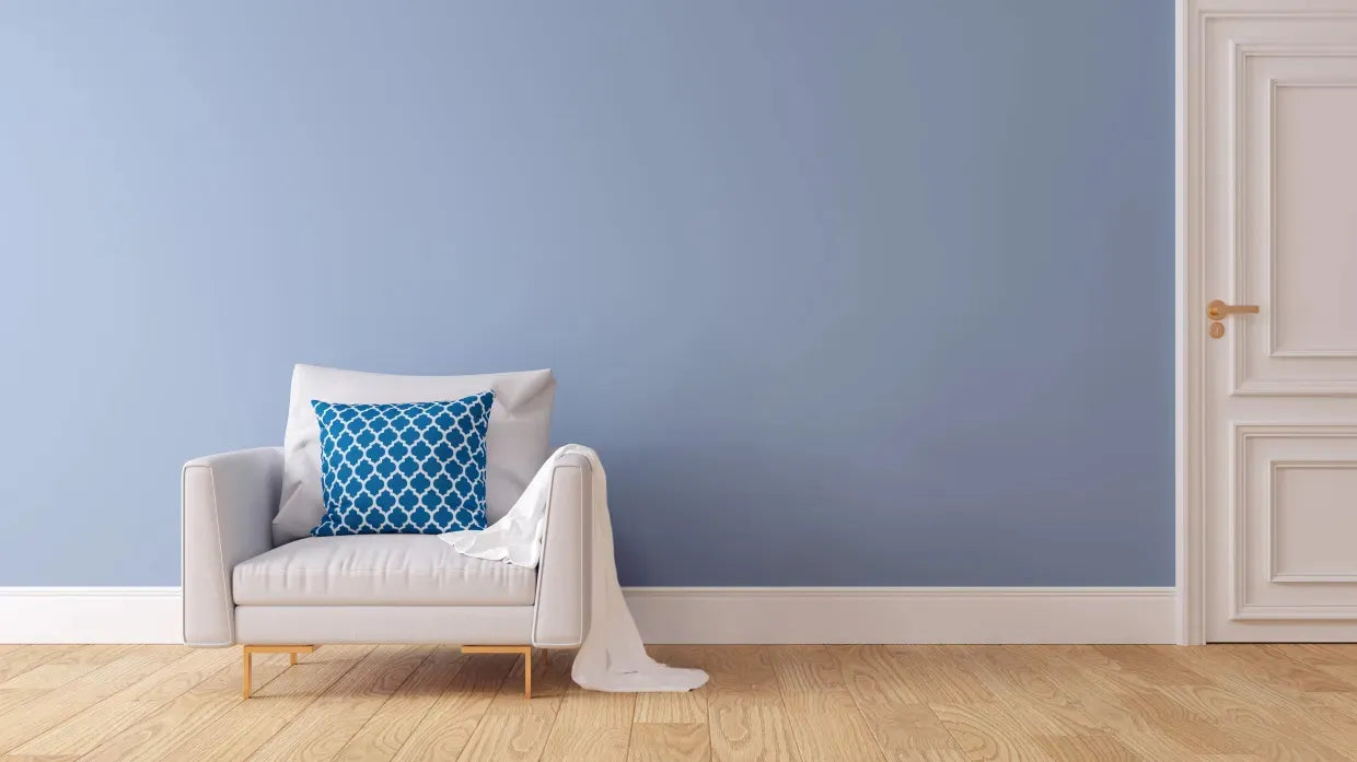 A cozy living room with a light gray armchair, accented by a blue patterned pillow and draped blanket, sits on a wooden floor. A small table holds a yellow clock and vase with daisies. The backdrop is a calming blue wall next to a white door, conveying a serene atmosphere.