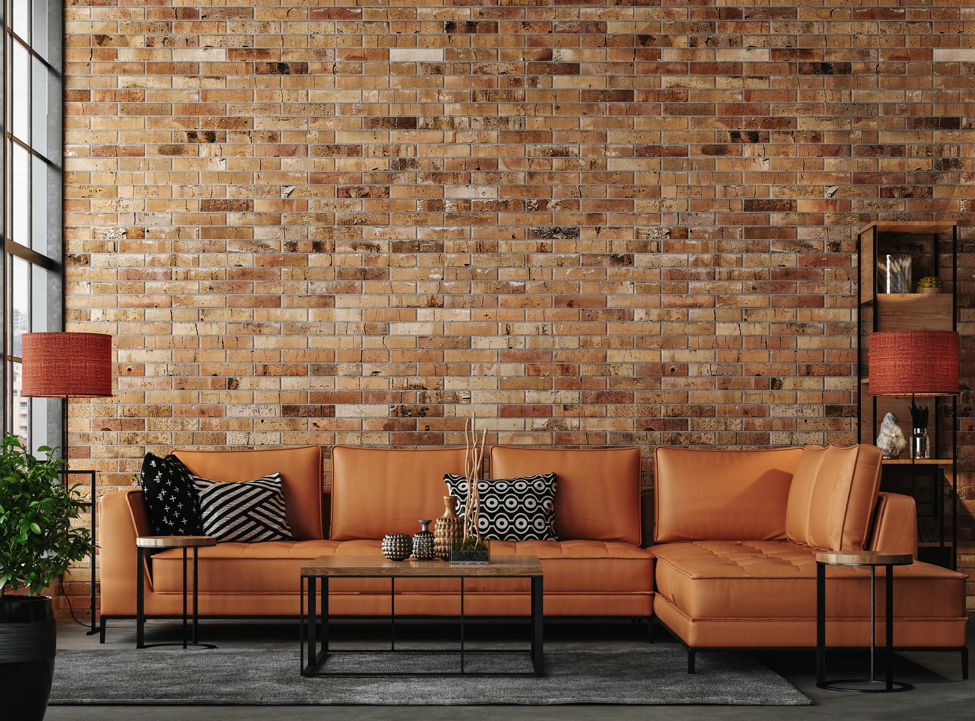 Stylish living room with a brown leather sectional sofa against a rustic brick wall. Features geometric pillows, modern tables, and red lamps, exuding a warm, contemporary vibe.