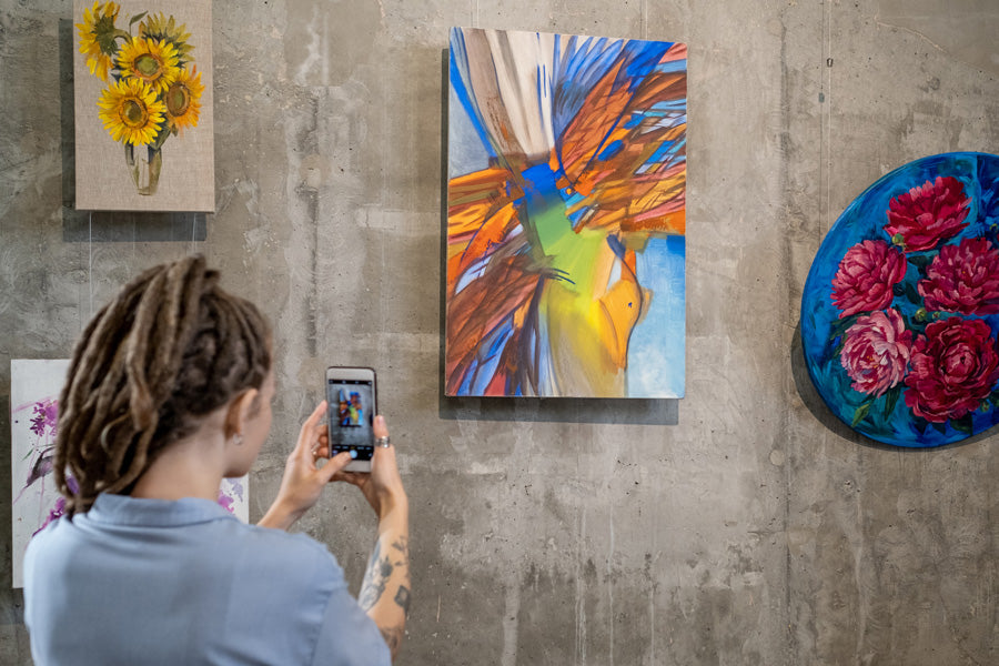 A person with dreadlocks photographs vibrant abstract and floral paintings on a concrete gallery wall, conveying creativity and inspiration.