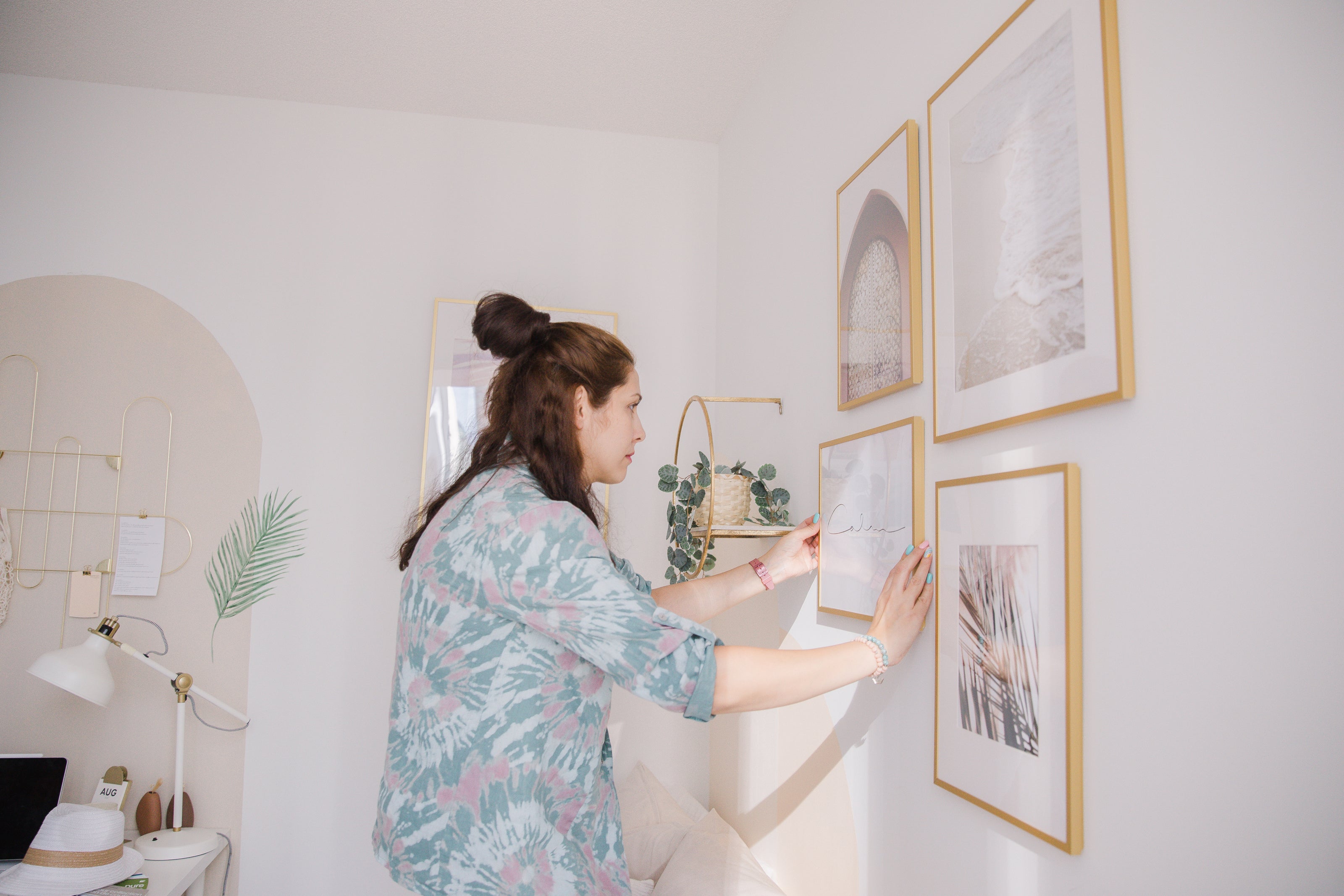 A woman in a floral shirt arranges a gallery of framed art on a white wall. The room is bright and tidy, with a serene and creative atmosphere.