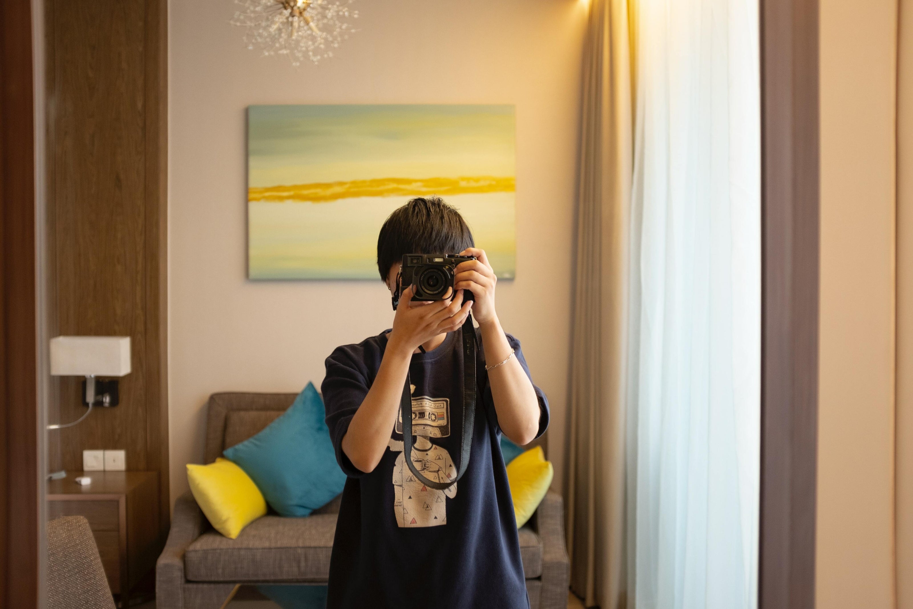 Person taking a mirror selfie in a cozy room, holding a camera. The room has a gray sofa with colorful cushions and a vibrant abstract painting.
