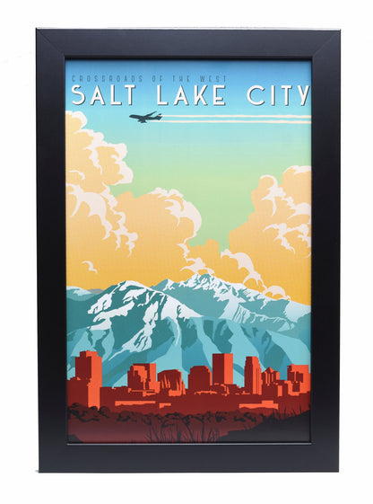 Framed poster of Salt Lake City skyline with red buildings, snow-capped mountains, and clouds in blue and orange sky. Plane flies overhead.